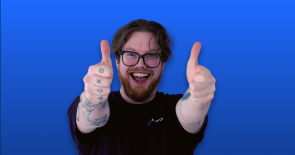 A bearded man with glasses gives two thumbs up and smiles excitedly against a blue background, showing tattoos on his arms.