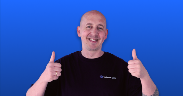 Smiling person giving a double thumbs-up, wearing a black "webinargeek" shirt, against a blue background.