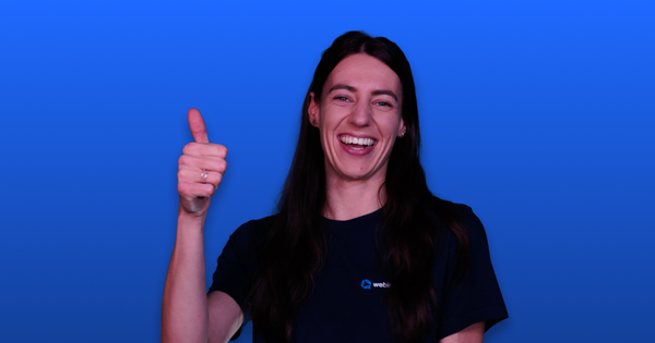 Person smiling widely, giving a thumbs-up against a blue background, wearing a dark shirt with a small logo.