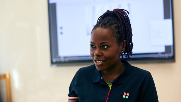Person with dark hair in a ponytail, wearing a navy polo shirt with colorful details, stands in front of a screen, engaged in conversation.