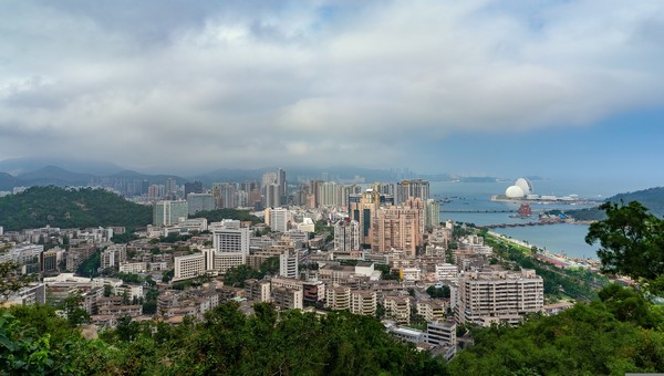 Aerial view of a coastal city with dense high-rise buildings, green hills, and a distinctive round white building by a bay under a cloudy sky.