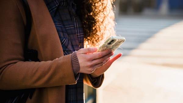 A person in a brown coat holds a smartphone with a floral case, standing outdoors in sunlight.