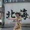 A person wearing a colorful floral outfit stands on a crosswalk, motioning forward, in front of a wall with large Chinese characters and "Be" visible.