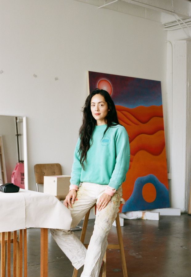 Camilla Engström sitting on a stool in her studio, blue and orange painting in the background