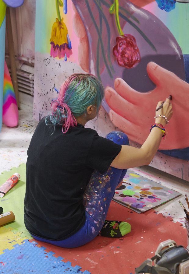 artist with blue and pink hair sitting on a foam mat, adding details to a large, colourful canvas
