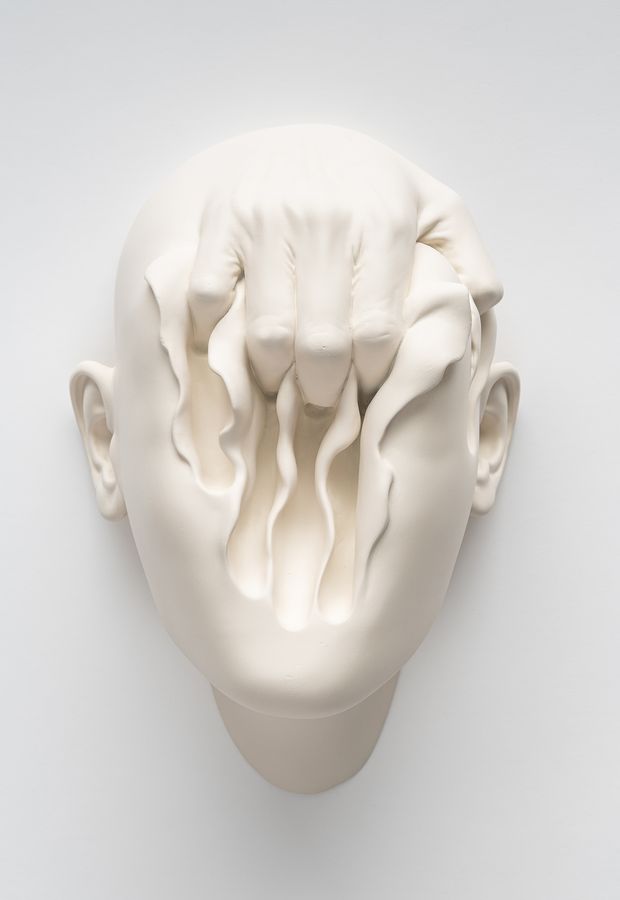 surreal, wall-mounted sculpture showing a hand digging deep into a face which seems to be made of soft clay
