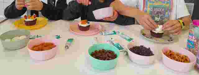Children decorating cupcakes with marshmallows, gummy bears, and sprinkles, surrounded by bowls of toppings like pretzels and chocolate chips.