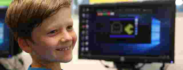 Smiling boy in a blue shirt sits in front of a computer displaying a pixelated game character on the screen.
