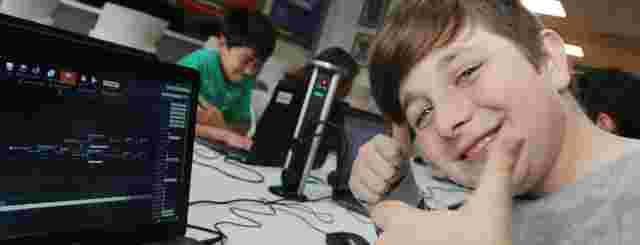 A smiling boy gives a thumbs-up next to a laptop displaying coding. Other children work on computers in the background.