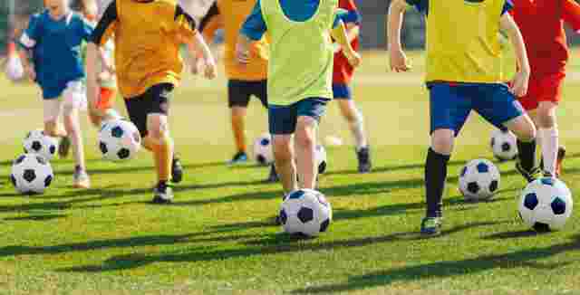 Children in brightly colored sportswear practicing soccer, each dribbling a ball on a green field.