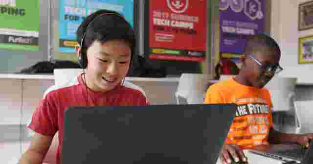 Two boys working on laptops in a tech camp classroom, one wearing headphones. Colorful camp posters are visible in the background.
