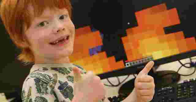 Child with red hair giving thumbs up in front of a computer screen displaying a pixelated game with fire graphics.