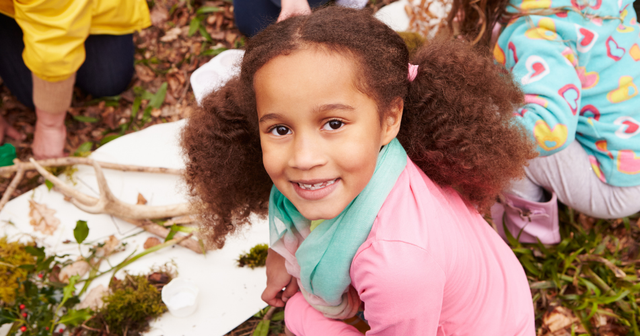 Girl with curly hair and pink shirt smiling while participating in an outdoor activity with other children and adults.