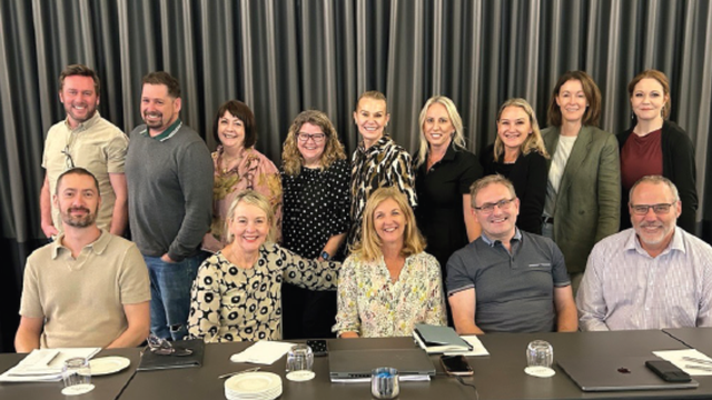 A group of people smiling, posing together in a conference room. They are seated and standing in two rows in front of a curtain.