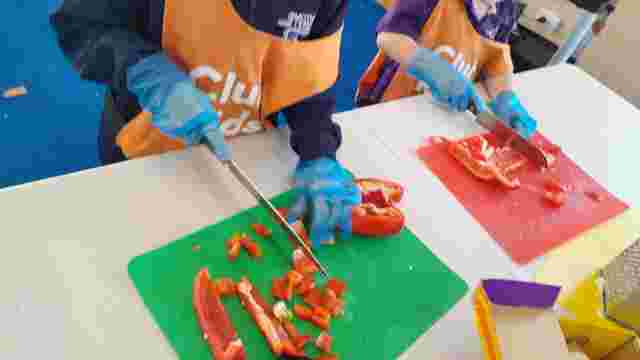 Children wearing aprons and gloves slice red bell peppers on colorful cutting boards in a cooking class.