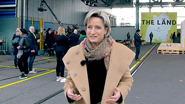 Eine Frau im Mantel steht im Vordergrund und redet. Im Hintergrund sieht man Kameras und ein großes "LÄND" Plakat.