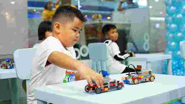 A child plays with a colorful toy car on a table, surrounded by other children in a bright, indoor setting with balloons in the background.