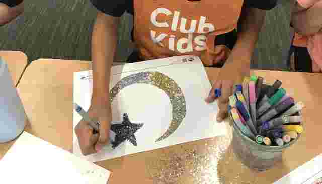 Child decorating a paper with a glittery crescent moon and star design, surrounded by markers and a cup on a classroom table.