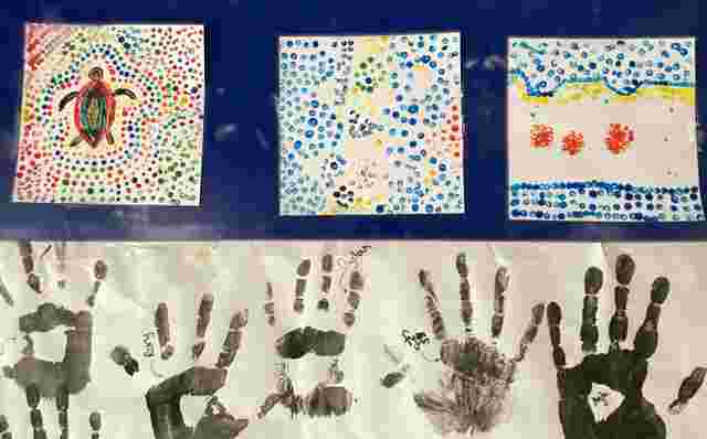 Three colorful dot paintings and multiple black handprints on paper displayed against a blue background.