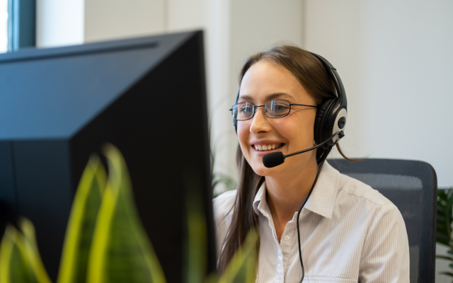 Woman with glasses and headset smiling while working at a computer in an office setting, with green plants in the foreground.