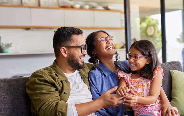 A happy family of three, including a man, woman, and young girl, sit on a couch laughing together in a modern living room.