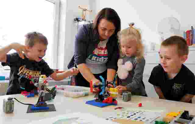 An adult helps three children build with colorful LEGO bricks on a table in a bright room.