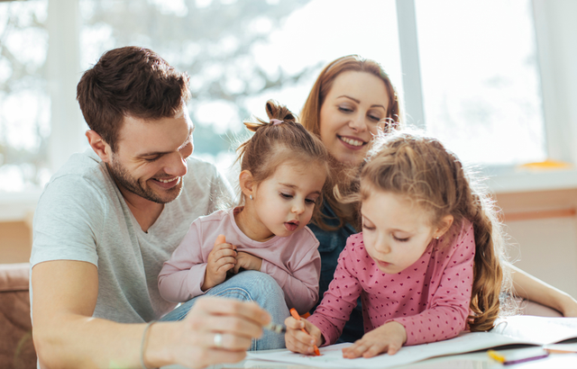 Family of four sitting together, smiling and engaged in drawing activities at home, with natural light streaming through a window in the background.
