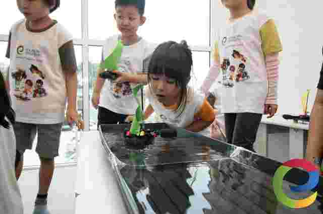 Children engaged in a science activity, testing small motorised LEGO sailboats in a water trough. They wear matching Young Engineers educational program shirts.
