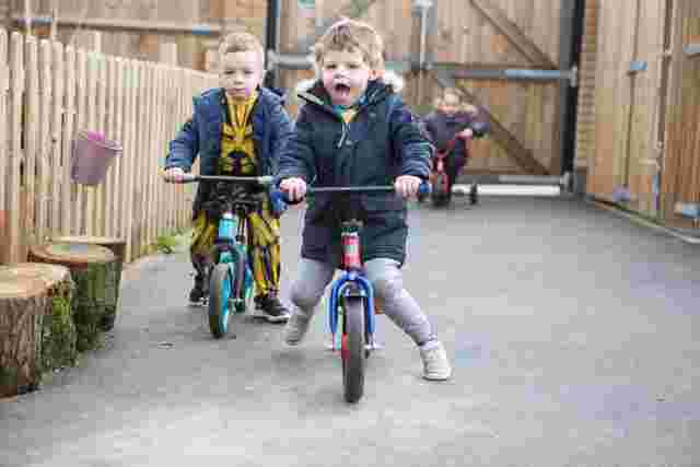 Two young children ride balance bikes energetically on a paved path, with a third child in the background. Wooden fence and gates are visible.