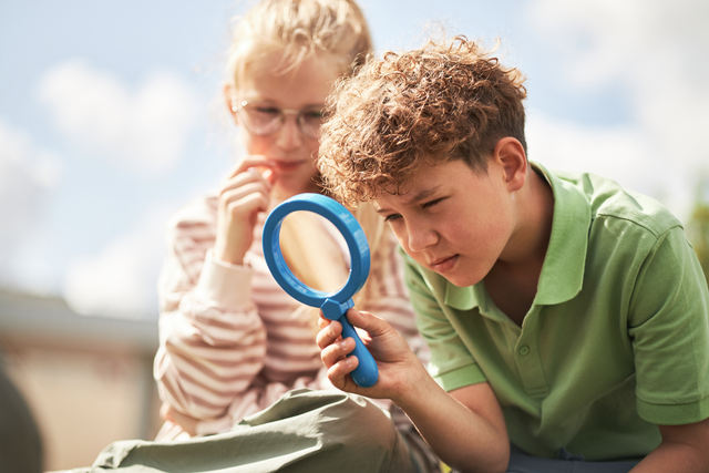 Two children examining something outdoors with a blue magnifying glass; one wears glasses and a striped shirt, the other in a green polo shirt.