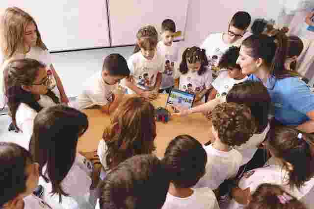 A group of children and a teacher gather around a table, looking at a tablet displaying colorful graphics in a classroom setting.