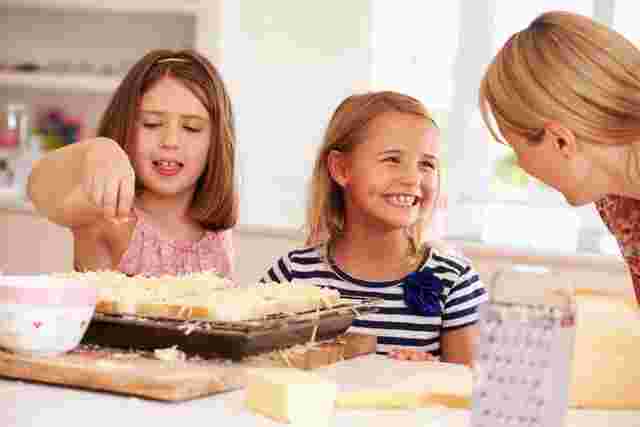 Two young girls and a woman in a bright kitchen, smiling and preparing cheesy bread on a tray.