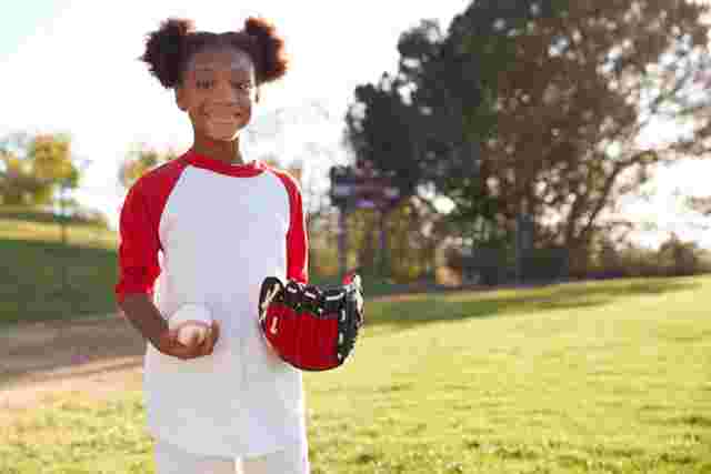 A smiling child in a baseball field, wearing a red and white shirt, holding a baseball and glove, standing on a sunny day.