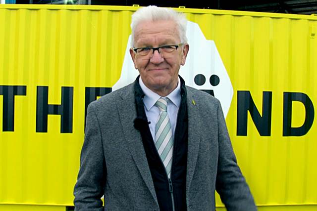 Ministerpräsident Winfried Kretschmann steht vor einem neongelben Container mit "THE LÄND" Schriftzug.