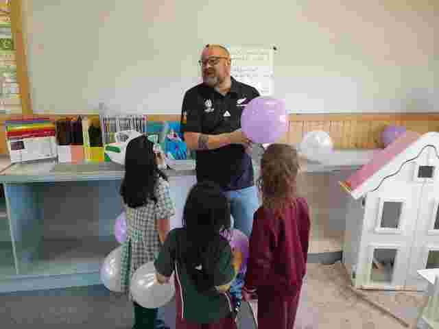 A teacher with purple balloons engages with three young girls in a classroom near a dollhouse.