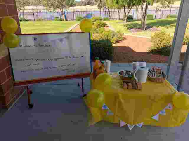 Outdoor table with a yellow tablecloth, drinks, snacks, and a sign reading "Happy R U OK? Day" decorated with yellow balloons.