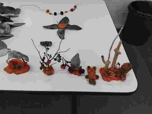 Natural craft made of leaves, twigs, pinecones, and orange clay on a white table, with a trash bin in the background.
