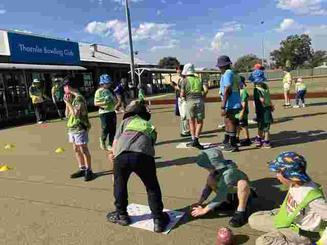 Children playing lawn bowls outside the Thornlie Bowling Club, interacting and wearing colorful hats under a sunny sky.