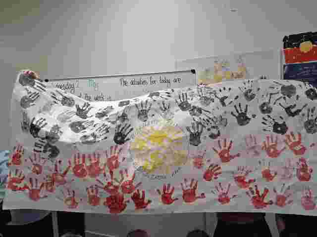 Banner with black, red, and yellow handprints, featuring "Reconciliation Week" text. Held in front of a classroom whiteboard.