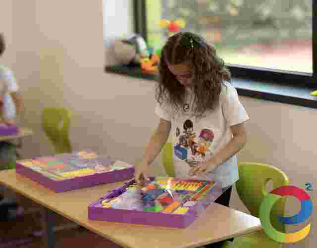 A young girl in a classroom assembles colorful building sticks from a kit on a desk. She wears a white Young Engineers T-shirt with cartoon characters.