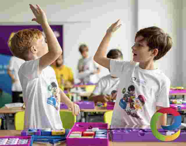 Two boys in matching Young Engineers T-shirts high-fiving in a classroom, surrounded by colorful educational kits and other children.