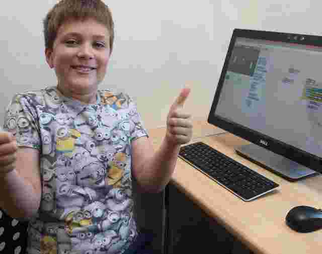 Boy smiling and giving thumbs up in front of a computer displaying a coding program. He is wearing a Minions-themed shirt.