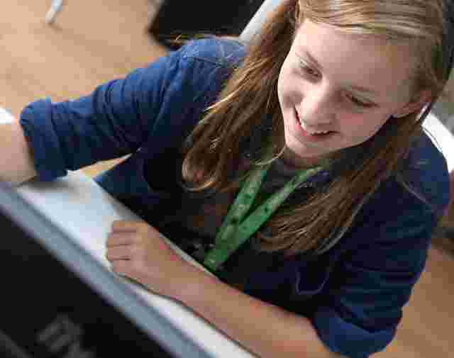 Smiling child with long hair, wearing a blue shirt and green lanyard, looks at a computer screen in a bright room.