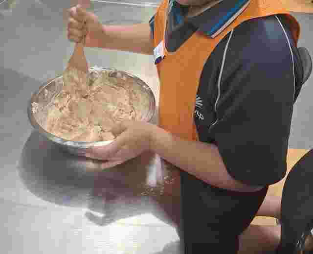 Child stirring batter in a metal bowl with a wooden spoon, wearing an orange vest and black shirt, on a stainless steel surface.