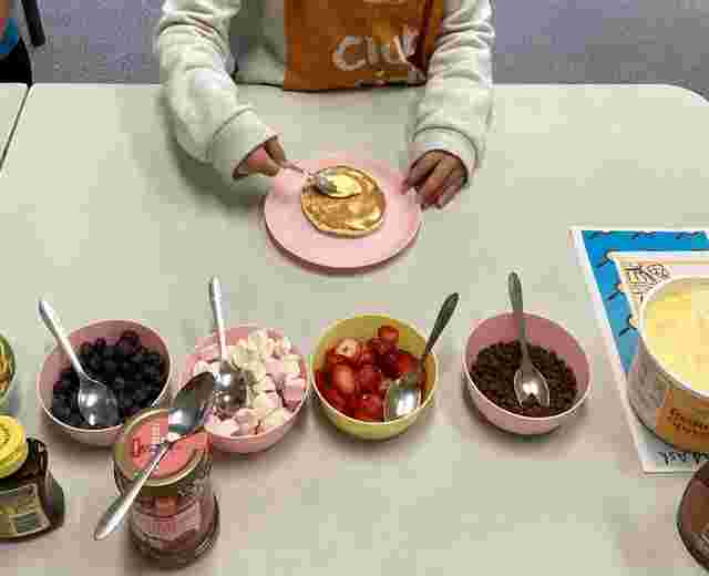Child spreading butter on a pancake at a table with bowls of blueberries, marshmallows, strawberries, chocolate chips, and syrup.