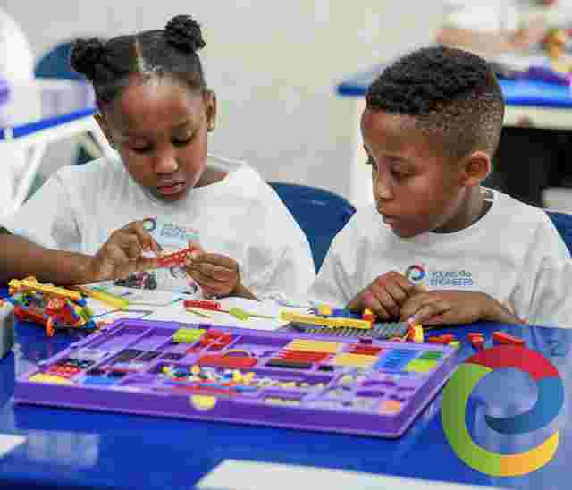 Two children in white shirts focus on building with colorful LEGO pieces at a table, surrounded by a purple tray of blocks.