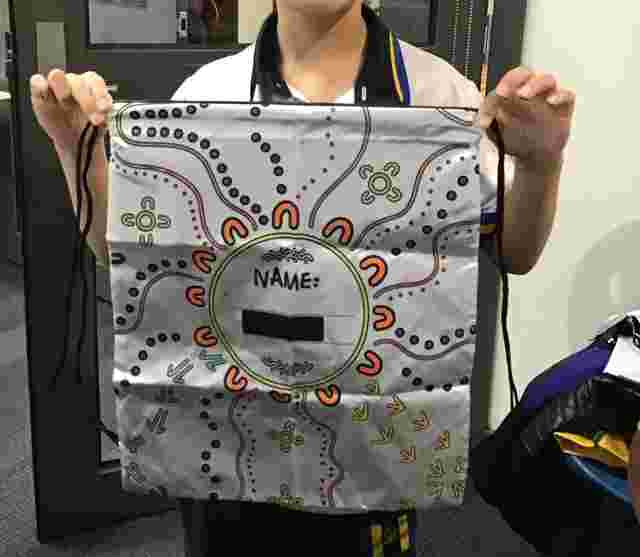 Person holding a drawstring bag decorated with colorful Aboriginal-style art and a blank "Name" label in the center.