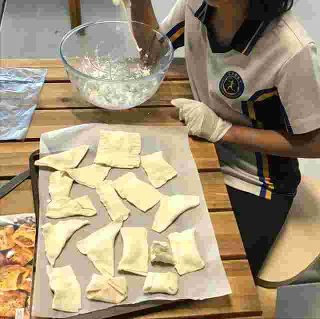 A person preparing pastry, shaping dough pieces on parchment paper, with a bowl of filling mixture on a wooden table.