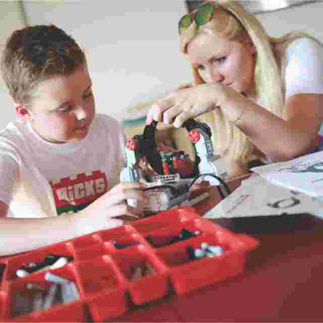 A boy and a woman work together on building a robot using a kit with red compartments on a table.