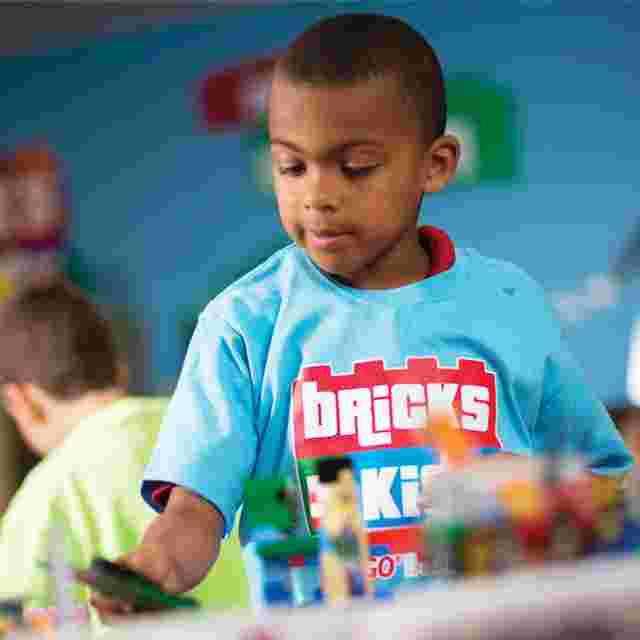 A child in a blue "Bricks 4 Kidz" shirt builds with colorful toy bricks in a play area.